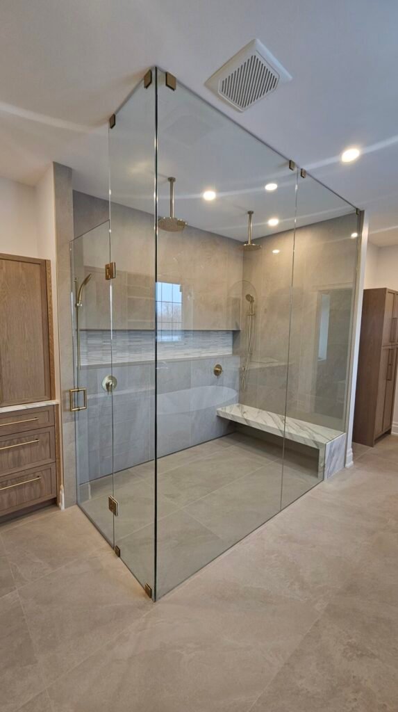 A custom glass shower and glass door, done in toronto, features gold hardware and beige tiles