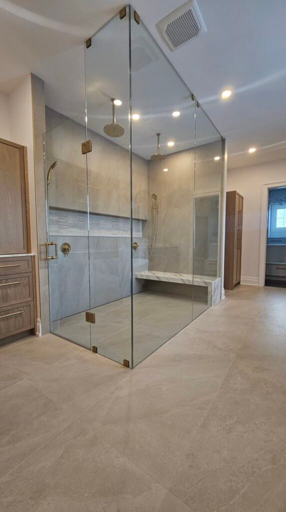 A custom glass shower and glass door, done in toronto, features gold hardware and beige tiles